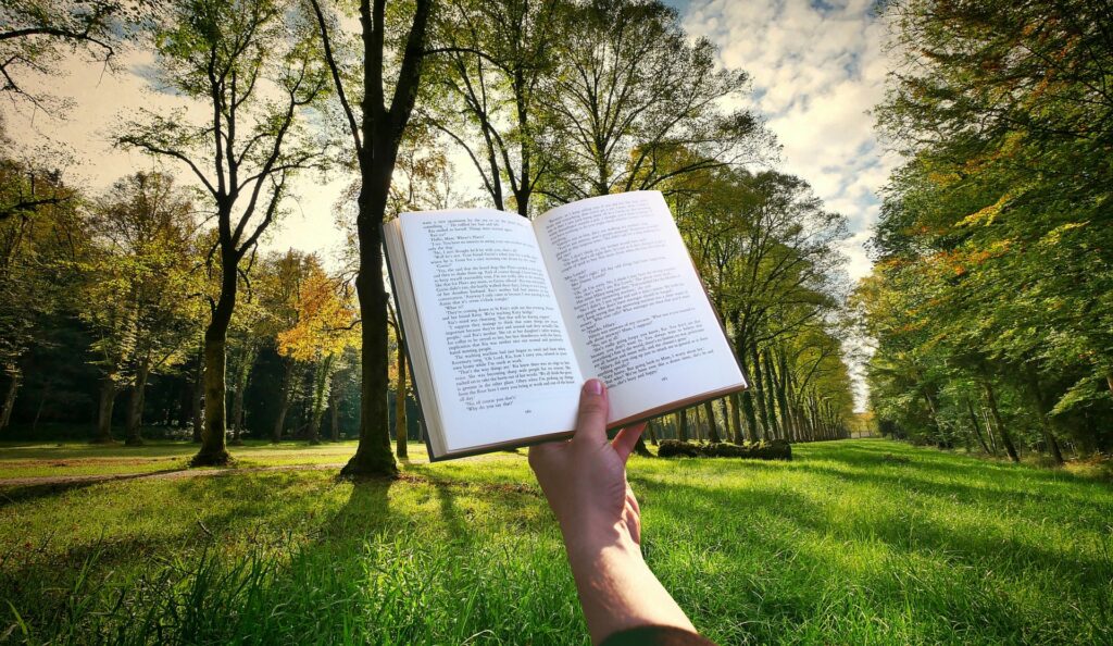 A hand holds an open book in the foreground. In the background you see many trees, green grass, and lots of sunlight.